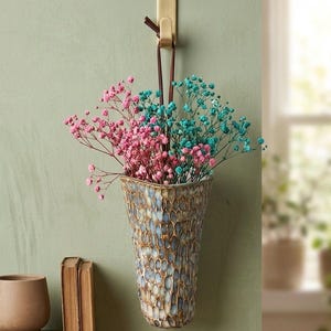 Peut inclure: Un vase mural avec un motif texturé bleu et beige, rempli de fleurs de gypsophile roses et turquoise. Le vase est suspendu à un crochet en laiton avec une lanière en cuir, ajoutant une touche de charme rustique.