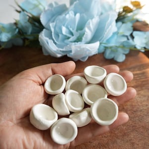 Puede incluir: Un conjunto de pequeños cuencos de cerámica blanca, cada uno con forma redondeada y acabado liso y brillante. Los cuencos se sostienen en una mano, con un fondo borroso de flores azules y una superficie de madera.