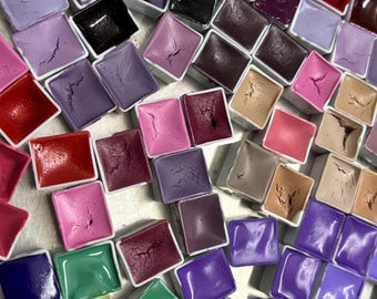 May include: A collection of watercolor paint pans in various colors, including shades of pink, purple, green, and blue. The paints are arranged in a grid pattern within small, square white containers. The colors range from light pastels to deep, rich hues.