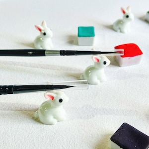 May include: White ceramic bunny figurines with black and silver paint brushes on a white surface. There are also small colorful blocks in the background.