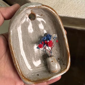 May include: A handmade ceramic dish with a small vase holding red, white, and blue dried flowers. The dish is oval-shaped with a hole at the top and a speckled, off-white glaze with brown edges. A unique decorative piece.