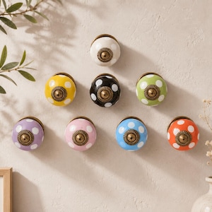 Può includere: Una collezione di otto pomelli rotondi in ceramica per mobili disposti a triangolo. Ogni pomello presenta un motivo a pois colorato con un fissaggio centrale in metallo color ottone anticato. I colori includono bianco, giallo, nero, verde, viola, rosa, blu e arancione.