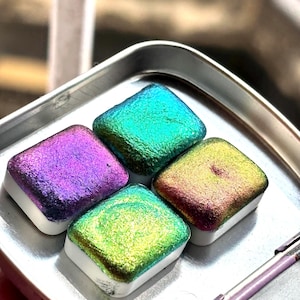 Könnte beinhalten: Vier quadratische Aquarellfarben in einer silbernen Schale. Die Farben haben ein schimmerndes, irisierendes Finish in Lila, Türkis, Grün und Gold. Jede Farbe befindet sich auf einer weißen Basis.