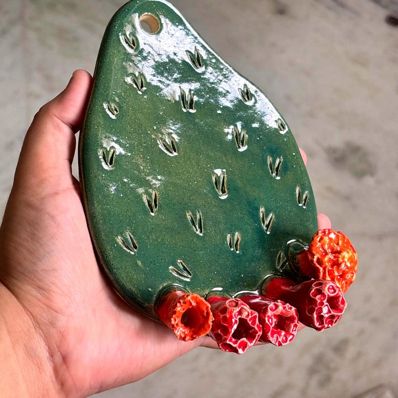 May include: A teardrop-shaped green ceramic wall hanging featuring a textured surface with small indentations and four bright orange and red tubular flower-like protrusions at the bottom. A circular hole is located at the top for hanging.