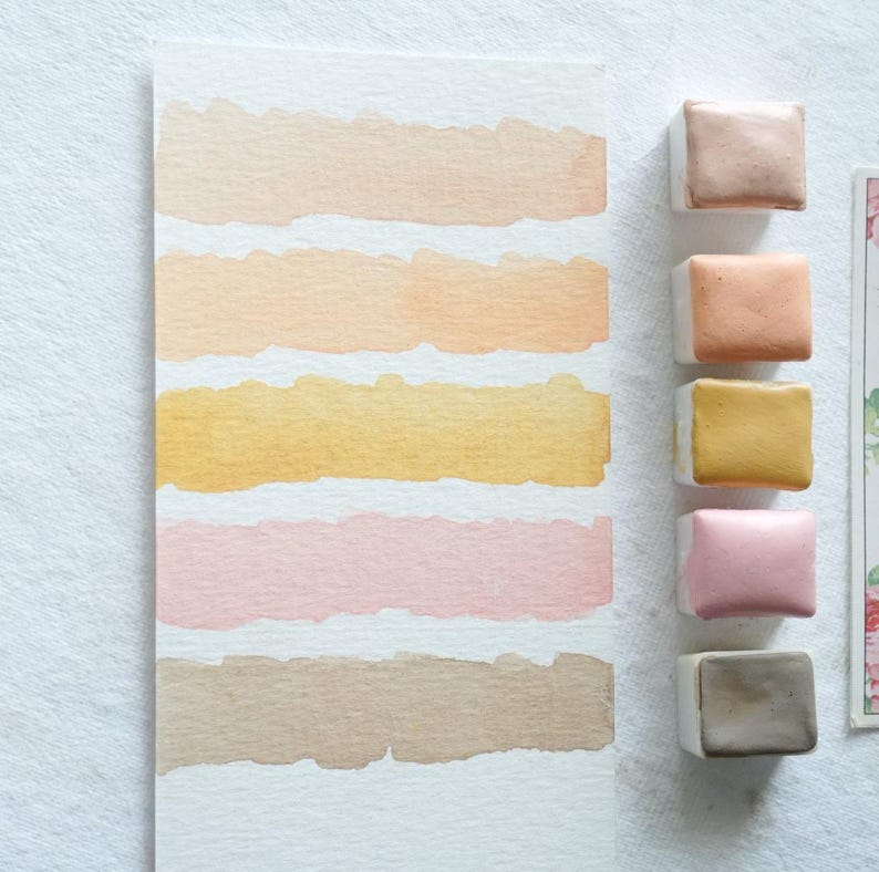 May include: A watercolor paint set featuring a palette of warm, earthy tones. The image shows a white paper with six horizontal brushstrokes in shades of beige, peach, and gold, alongside five square paint cakes in matching colors.