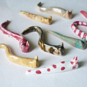 Op de afbeelding: Een verzameling keramische katvormige eetstokjessteunen in verschillende kleuren en patronen. De collectie bevat stukken in roze, geel, bruin en wit met rode stippen. Elk stuk is ongeveer 10 cm lang.