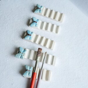 Puede incluir: Soportes para pinceles de cer&aacute;mica blanquecina con un dise&ntilde;o ondulado, cada uno coronado con un acento de mariposa azul claro. Dos pinceles descansan en los soportes. Los soportes est&aacute;n sobre una superficie blanca.