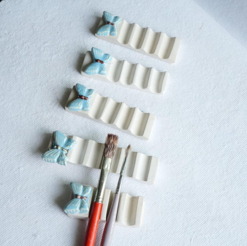 Puede incluir: Soportes para pinceles de cer&aacute;mica blanquecina con un dise&ntilde;o ondulado, cada uno coronado con un acento de mariposa azul claro. Dos pinceles descansan en los soportes. Los soportes est&aacute;n sobre una superficie blanca.