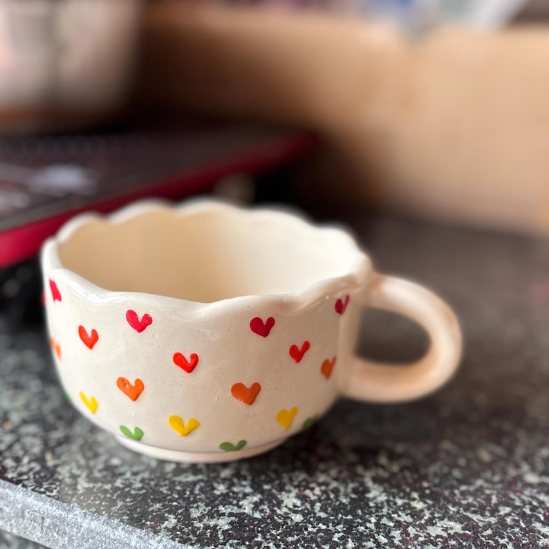 May include: A cream-colored ceramic mug with a scalloped rim and a handle. The mug is decorated with small, colorful hearts in red, orange, yellow, and green. The mug is sitting on a dark surface.