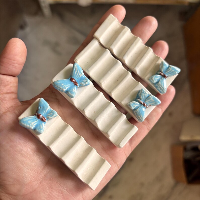 Puede incluir: Juego de cuatro reposautensilios de cer&aacute;mica. Cada uno es de color blanco roto con un dise&ntilde;o estriado y una mariposa azul claro con detalles marrones. Ideales para la cocina.