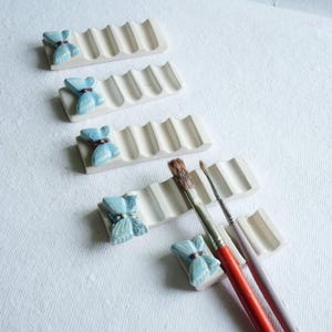 Puede incluir: Cuatro soportes para pinceles de cer&aacute;mica blanquecina con un dise&ntilde;o ondulado. Cada soporte tiene un adorno de lazo azul claro con un centro marr&oacute;n. Dos pinceles descansan en un soporte. Los soportes est&aacute;n sobre una superficie blanca.