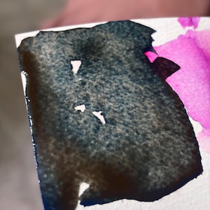Könnte beinhalten: Nahaufnahme eines Aquarellgemäldes mit dunkelblauen und braunen Farbtönen, die in ein leuchtendes Pink übergehen. Die Farbe wird auf weißes Papier aufgetragen, mit sichtbaren Pinselstrichen und einer strukturierten Oberfläche. Das Kunstwerk zeigt eine Mischung aus Farben.
