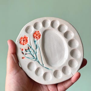 Op de afbeelding: Een ronde, gebroken witte keramische verfpalet met een centrale mengbak omringd door twaalf kleine cirkelvormige inkepingen. Het oppervlak is versierd met een handgeschilderd ontwerp van twee oranje bloemen met groene stengels en bladeren.