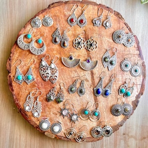 May include: A collection of silver-toned earrings with various designs, including turquoise, red, and purple gemstones, displayed on a wooden surface. The earrings feature intricate filigree, geometric shapes, and fan-like structures, showcasing a bohemian aesthetic.