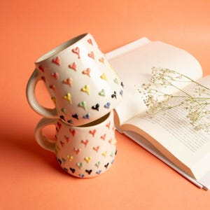 May include: Two stacked ceramic mugs with a cream-colored base, adorned with raised, colorful heart-shaped designs. The hearts are in various colors, including red, yellow, green, blue, and black. A book with open pages and a sprig of white flowers are in the background.