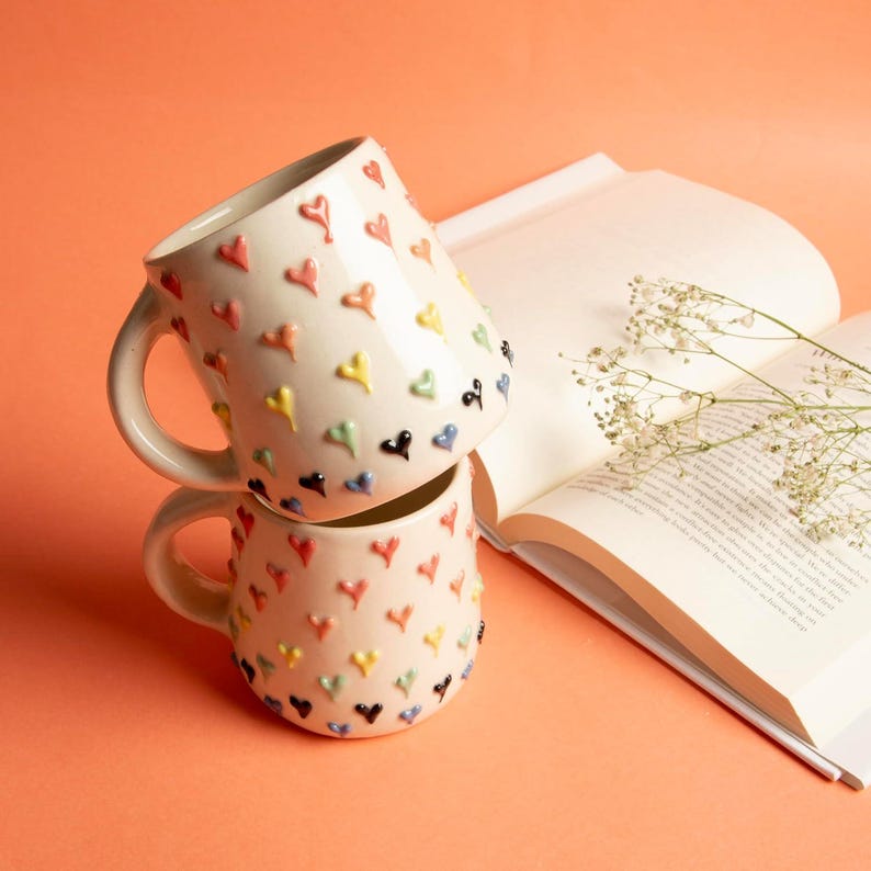 May include: Two stacked ceramic mugs with a cream-colored base, adorned with raised, colorful heart-shaped designs. The hearts are in various colors, including red, yellow, green, blue, and black. A book with open pages and a sprig of white flowers are in the background.