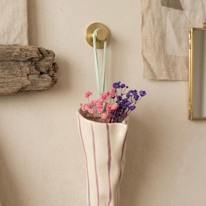 May include: A wall-mounted ceramic vase with an irregular, organic shape and vertical purple stripes. The vase is suspended by a light green strap from a circular brass hook and contains a small bouquet of dried pink and purple flowers.