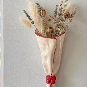 Peut inclure: Un vase mural en céramique couleur crème en forme de bouquet enveloppé, avec un bord peint en rouge et un nœud rouge sculpté à la base. Le vase contient un arrangement de graminées séchées, de brins de lavande et de petites gousses brunes.