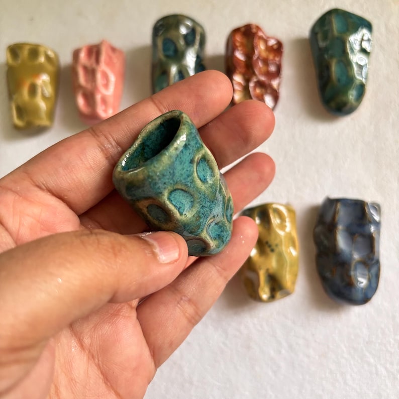May include: A collection of small, handmade ceramic vases in various colors, including teal, pink, yellow, and blue. The vases have a textured surface and a unique, organic shape. One teal vase is held in a hand.