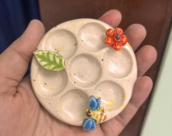 Puede incluir: Una paleta de pintura de cerámica blanca con seis pozos redondos. La paleta está decorada con una hoja verde, una flor roja y una abeja azul y amarilla.
