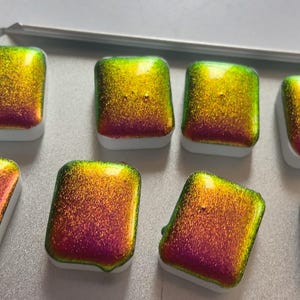 K&ouml;nnte beinhalten: Acht schimmernde Aquarellfarben in einer Metallbox. Die Farben wechseln von Gr&uuml;n &uuml;ber Gold zu Rot. Jedes rechteckige Feld sitzt auf einem wei&szlig;en Sockel. Ideal f&uuml;r K&uuml;nstler.