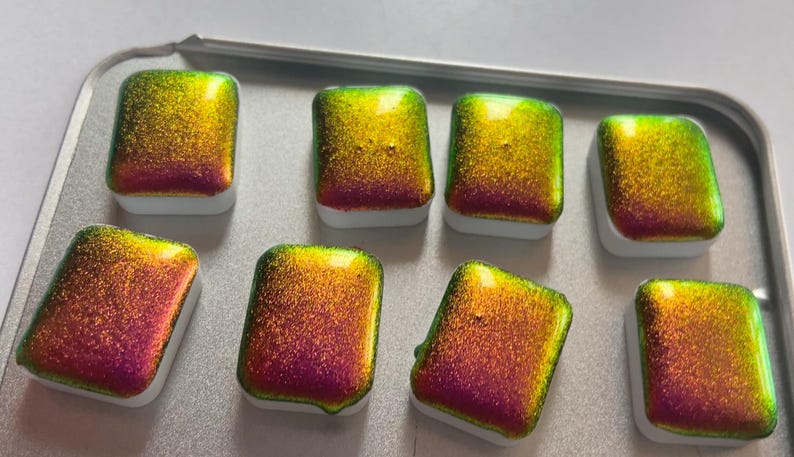K&ouml;nnte beinhalten: Acht schimmernde Aquarellfarben in einer Metallbox. Die Farben wechseln von Gr&uuml;n &uuml;ber Gold zu Rot. Jedes rechteckige Feld sitzt auf einem wei&szlig;en Sockel. Ideal f&uuml;r K&uuml;nstler.