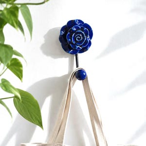 May include: A decorative wall hook with a blue ceramic rose design. The hook is holding a beige tote bag. The rose has detailed petals and a glossy finish. The hook is mounted on a white wall, with a green plant in the background.