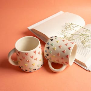 May include: Two cream-colored ceramic mugs with heart-shaped embellishments in various colors. The mugs have a rounded shape with a handle and are placed on a peach-colored surface. A book is open in the background.