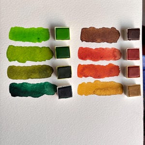 May include: A collection of watercolor paints in various shades of green, brown, orange, and yellow. Each color is displayed as a swatch and in a small square pan. The swatches are on a white surface.