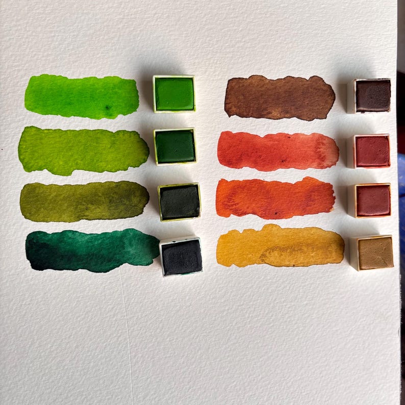 May include: A collection of watercolor paints in various shades of green, brown, orange, and yellow. Each color is displayed as a swatch and in a small square pan. The swatches are on a white surface.