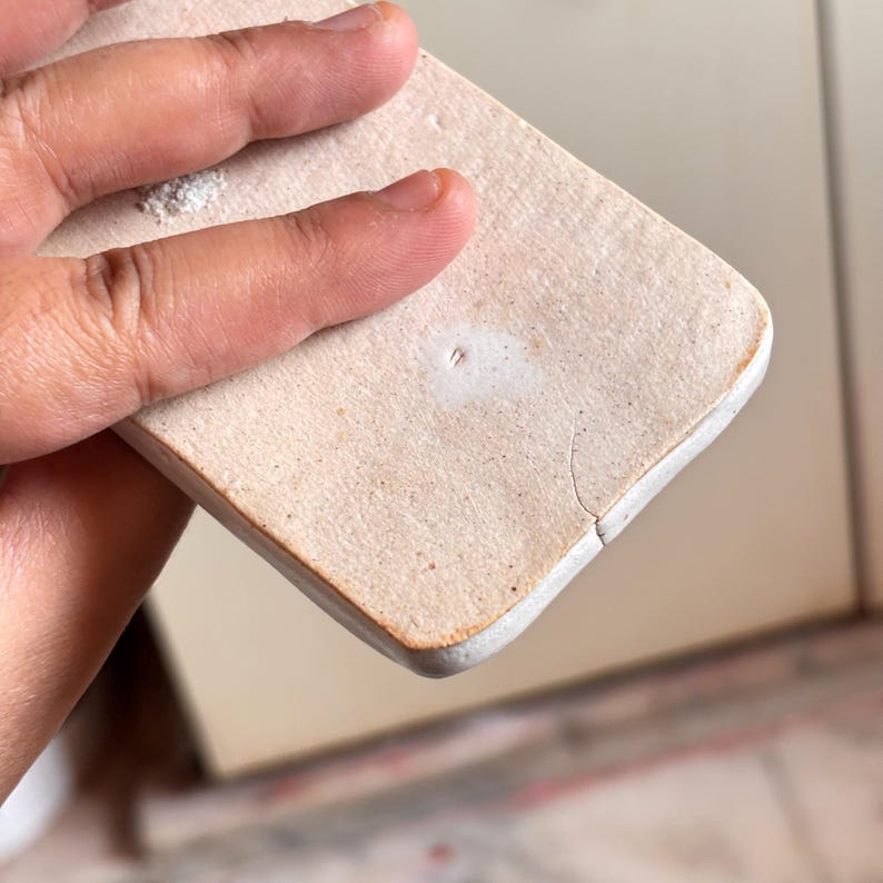 Pu&ograve; includere: Un sottobicchiere rettangolare in ceramica bianco sporco con angoli arrotondati. Il sottobicchiere ha una superficie leggermente strutturata e una crepa visibile lungo un bordo. &Egrave; tenuto in mano.