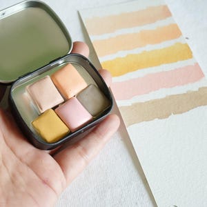 May include: A small, open metal tin containing six watercolor paint squares in shades of yellow, pink, peach, and brown. A watercolor swatch card with horizontal stripes of similar colors is next to the tin. The tin is held in a hand.