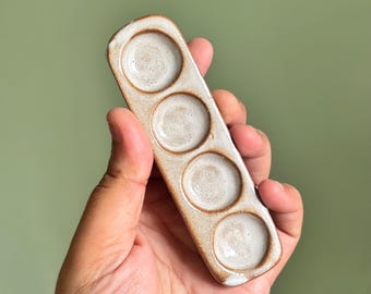 May include: A rectangular ceramic paint palette featuring four circular wells arranged in a single vertical row. The palette has a speckled beige and brown exterior with a smooth, glossy white glaze finish inside each of the four circular indentations.