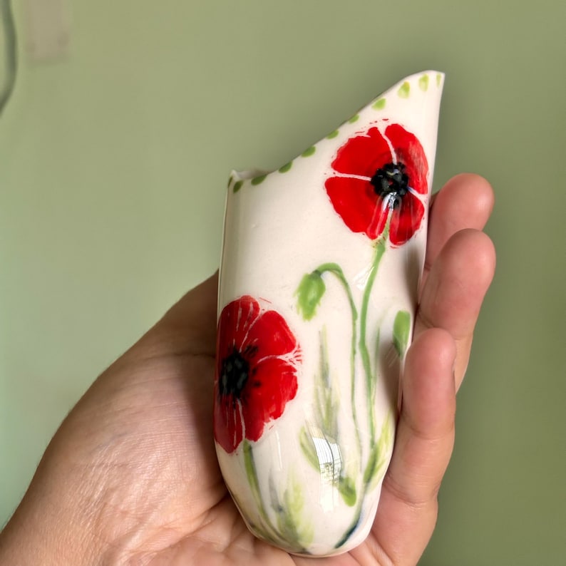 Może przedstawiać: Mały, kremowy wazon ceramiczny lub wazon ścienny z ręcznie malowanymi czerwonymi makami z czarnymi środkami i zielonymi łodygami. G&oacute;rna krawędź jest ukośna i ozdobiona serią małych zielonych kropek wzdłuż obrzeża przedmiotu.