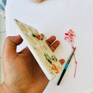 May include: White ceramic paint palette with 16 wells, decorated with green leaves and orange flowers. The palette is held in a hand.