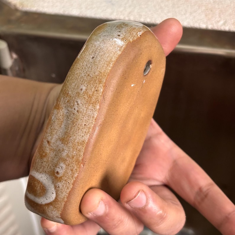 May include: A handmade, rectangular ceramic soap dish with a hole for drainage. The dish is a warm brown color with a white and tan glazed edge. The soap dish is held in a hand.