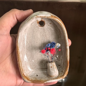 May include: A handmade ceramic wall pocket with a small vase holding red, white, and blue dried flowers. The pocket is a light gray color with a speckled texture and a brown rim. It has a hole at the top for hanging.