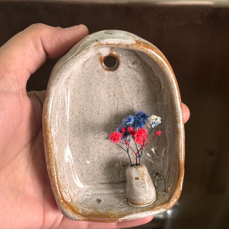 May include: A handmade ceramic wall pocket with a small vase holding red, white, and blue dried flowers. The pocket is a light gray color with a speckled texture and a brown rim. It has a hole at the top for hanging.