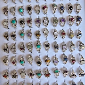 May include: A collection of 60 silver rings with various gemstone settings. The rings feature a delicate, ornate design and are arranged in rows on a white display board.