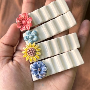 May include: Four white ceramic rectangular tiles with raised ridges. Each tile features a different colourful flower design: a red flower, a blue flower, a yellow flower, and a purple flower.