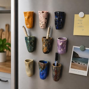 Peut inclure: Neuf petits gobelets en céramique aux formes irrégulières fixés sur une surface de réfrigérateur. Les gobelets présentent divers émaux, notamment orange, rose tacheté, bleu foncé, vert, marron, violet, beige et multicolore. Trois contiennent des stylos.