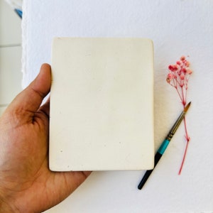 May include: A white ceramic square tile with a smooth, slightly textured surface. The tile is being held by a hand.