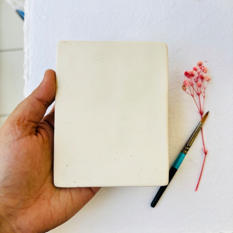 May include: A white ceramic square tile with a smooth, slightly textured surface. The tile is being held by a hand.
