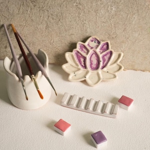 Puede incluir: Una paleta de pintura de cerámica blanca con forma de flor de loto con detalles en morado. La paleta está rodeada por un portapinceles de cerámica blanca con tres pinceles dentro y tres pequeños cuadrados de pintura de acuarela.