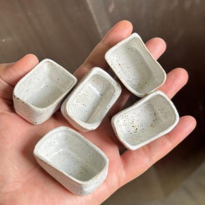 May include: Five small, rectangular ceramic vessels with a speckled off-white glaze. Each miniature container features rounded corners and a smooth, hollow interior, showcasing a consistent artisanal finish across all five individual pieces.