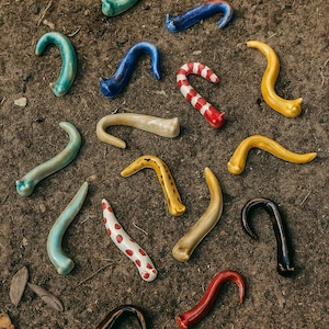 May include: A selection of colourful, curved ceramic hooks in various shades and patterns. The hooks are arranged on a brown, earthy surface. Colours include blue, red, yellow, and white, with some featuring stripes or polka dots.