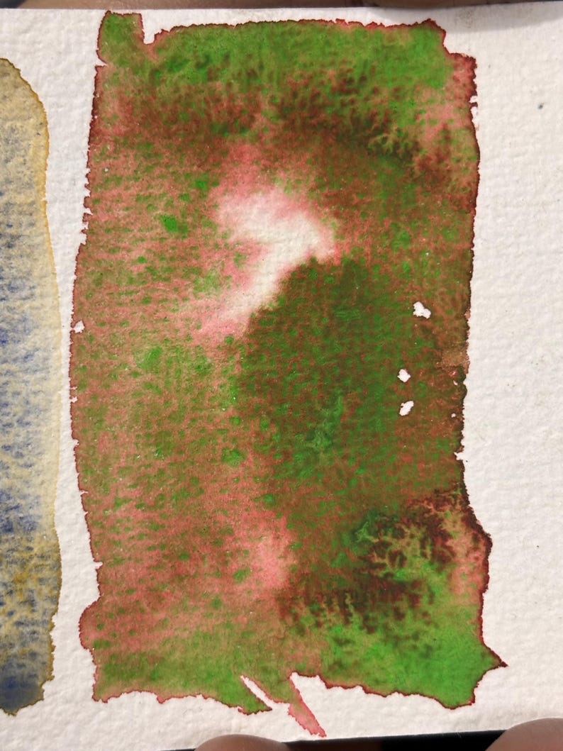 May include: A rectangular watercolor swatch with a blend of green, red, and white hues. The colors are applied with a textured effect, creating a mottled appearance. The edges of the paper are visible.