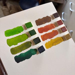 May include: Watercolor paint set with various colors, including greens, browns, oranges, and yellows. Each color is displayed as a swatch next to its corresponding paint pan. The paints are on a white paper surface.