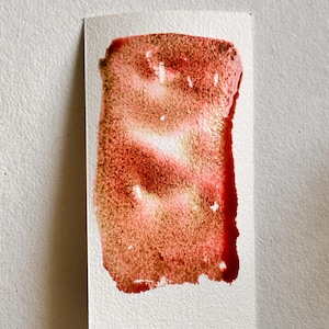 May include: A rectangular watercolor swatch on white paper. The paint is a blend of red, pink, and brown hues, with a textured appearance. The edges of the paint are slightly uneven, and the colors blend together.