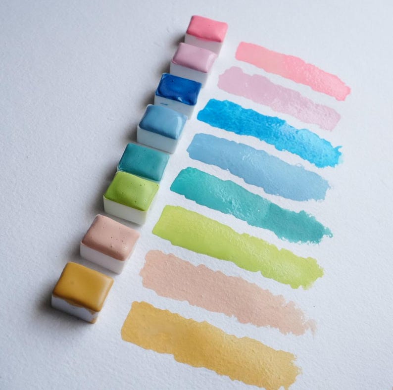 May include: A collection of watercolor paint pans in various colors, including pink, blue, green, and yellow. Each pan is next to a corresponding color swatch on white paper, showcasing the paint's hue and texture. Ideal for artists.